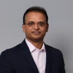 Man in suit with glasses, neutral background.