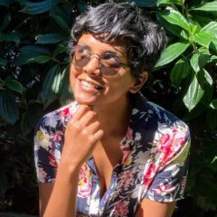 A woman with short hair and glasses smiling.|A woman smiling for the camera in front of some bushes.|A woman smiling for the camera in front of some bushes.