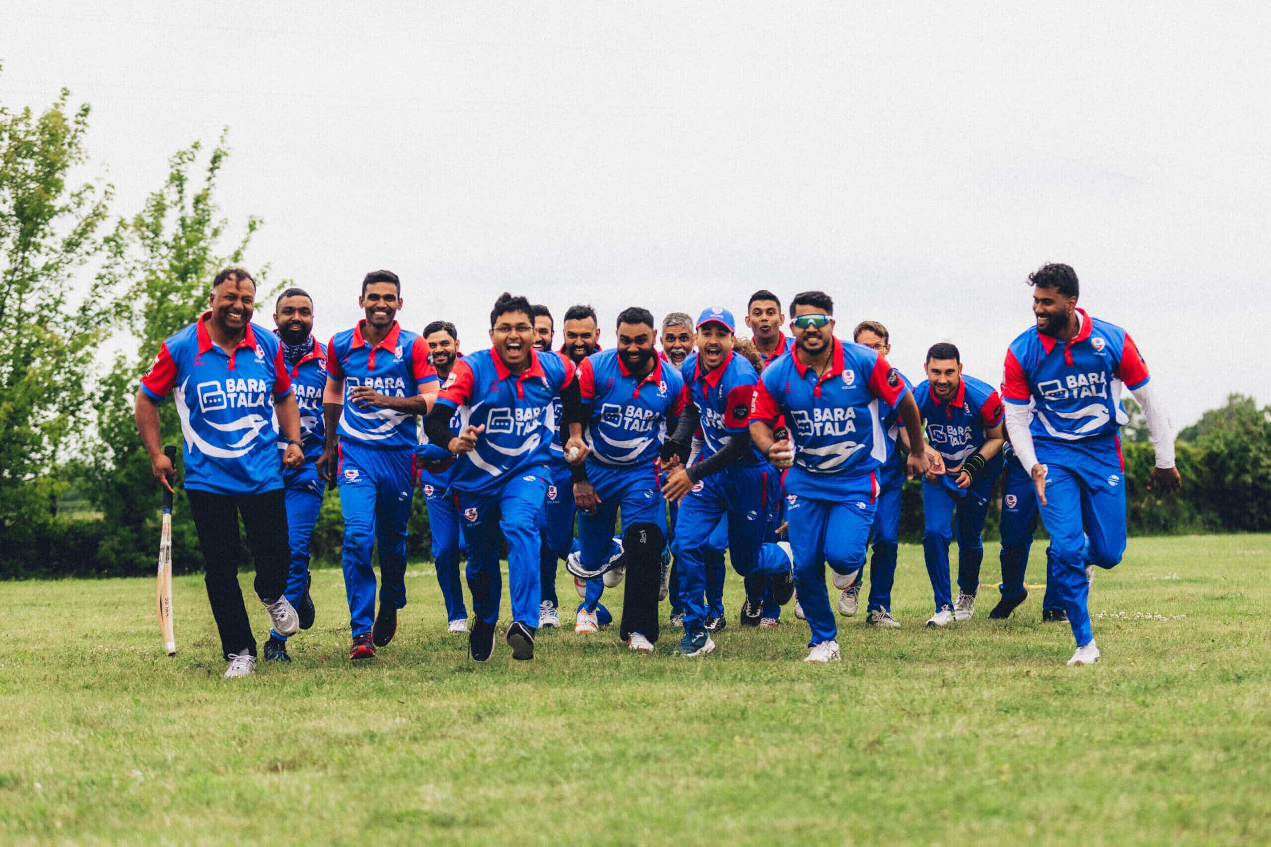Cricket team running on grassy field.|Cricket team posing together on grass field.|Smiling man in blue cricket uniform outdoors.