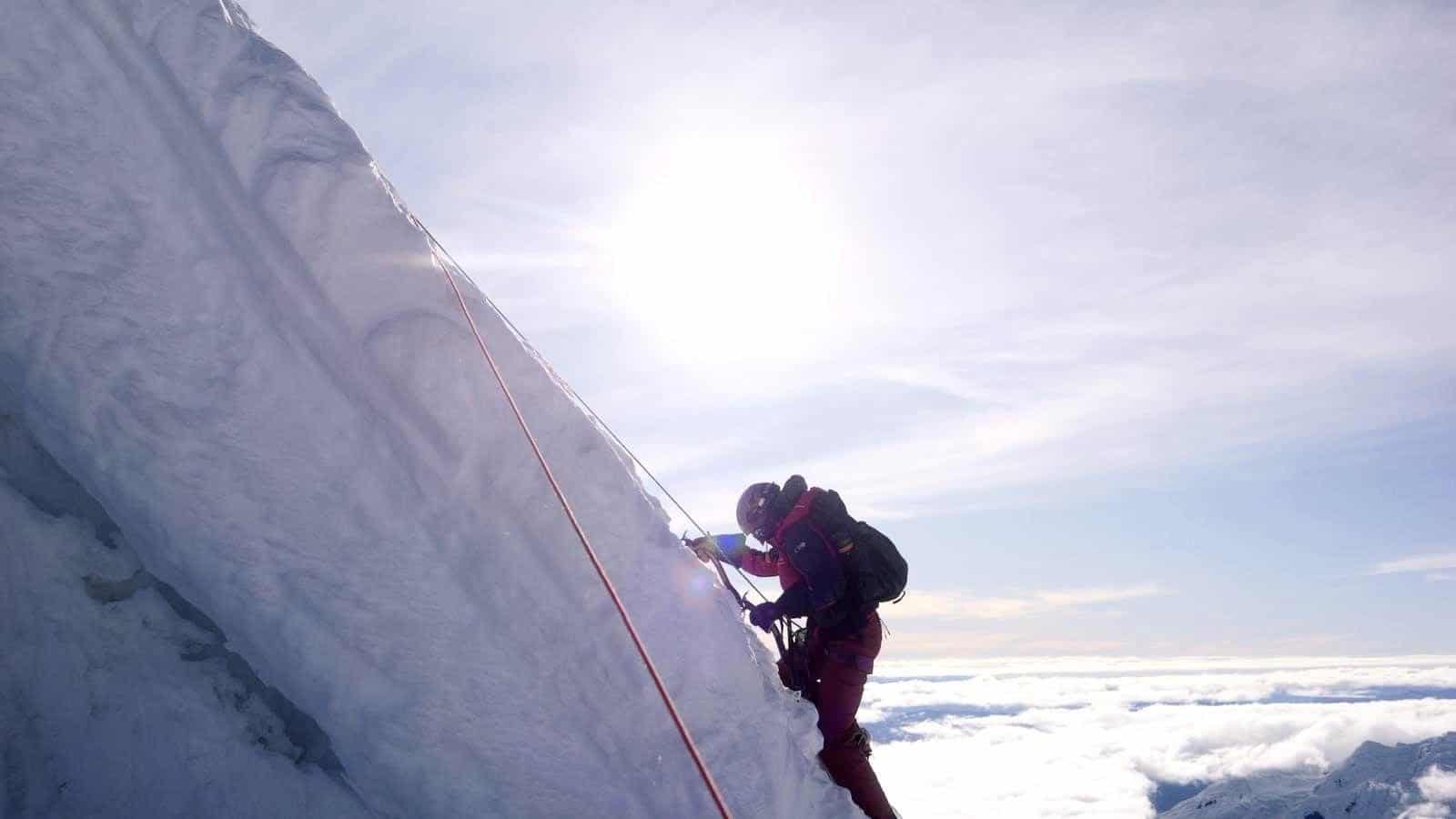 Climber ascending steep snowy slope with rope|Person wearing blue goggles and patterned face covering|Person wearing blue reflective goggles and bandana|Smiling woman resting chin on hand