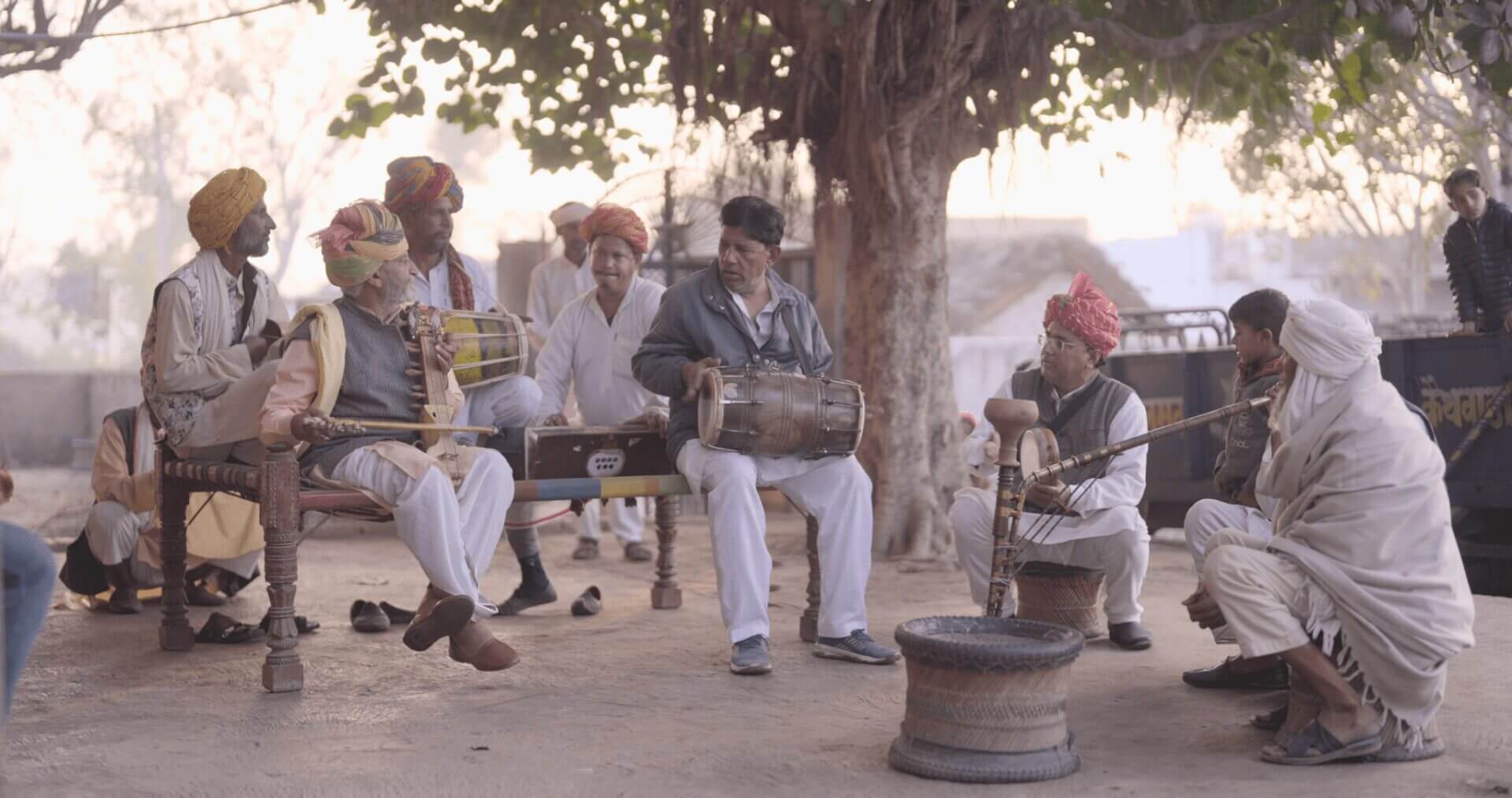 Rural Indian musicians playing traditional instruments|Young woman with long hair in yellow top|Seated man beside Indian flag photographed outdoors|Rajasthani folk musicians performing outdoors|Two people in white clothing walking through field