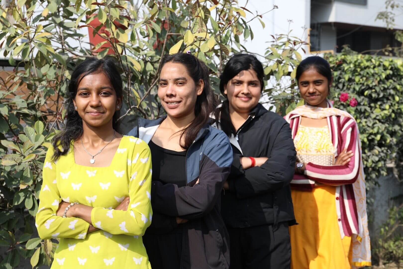 Four women standing, smiling, arms crossed outside.|Breakthrough logo against gender-based violence.|Person smiling and sitting outdoors, surrounded by greenery.|Group walking on a sunny street.|Young girl speaking at outdoor event.|Girl speaking at community event outdoors.|Group of four people smiling on bench.|School event with students on stage.