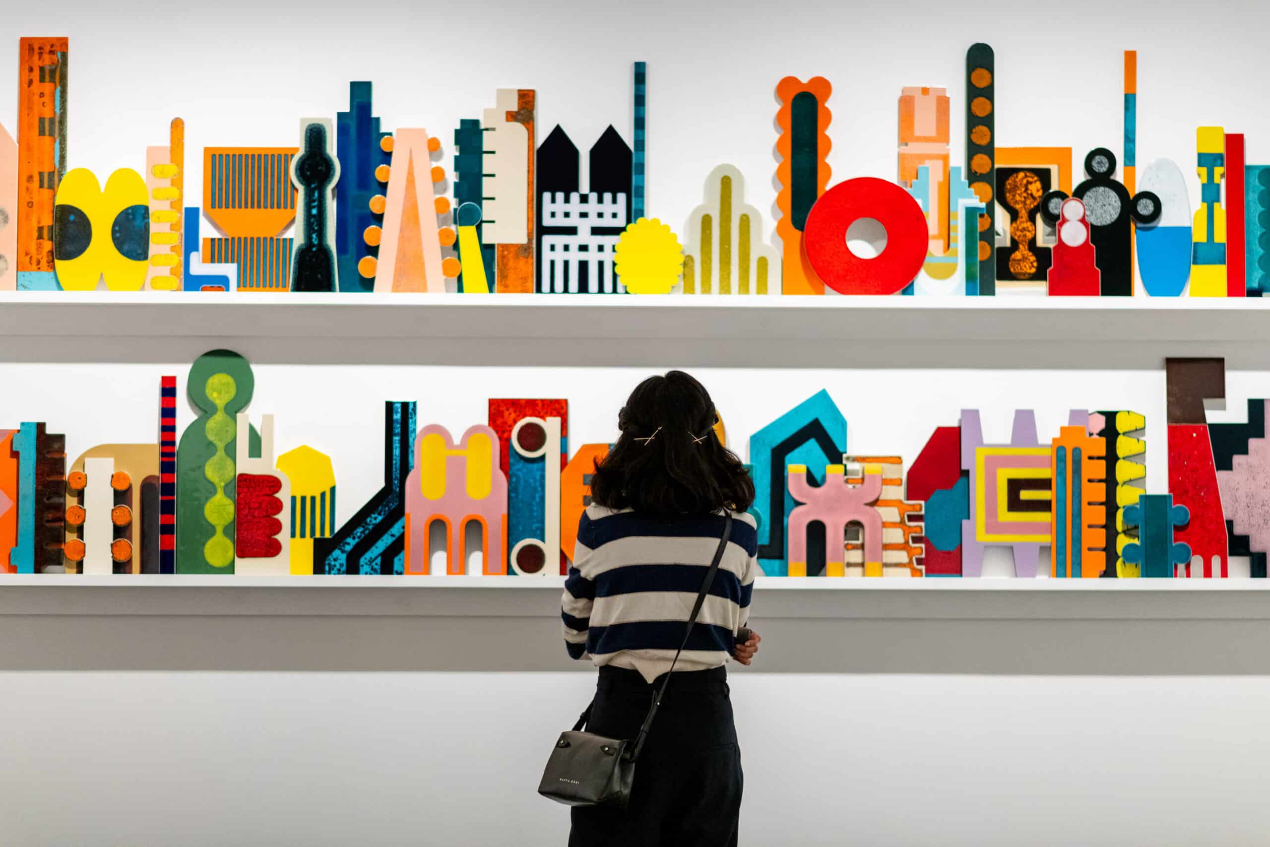 Person viewing colorful abstract art installation on shelves|Two children seated watching colorful projected illustration|Two people reading inside orange circular pod|Three children on mini trampolines viewing art|||Woman standing before blue painting in gallery|Professional headshot of man with gray hair|Museum display of antique wooden wagon wheels|Colorful decorated vintage vehicles displayed in museum|Colorful decorated tuk-tuk and rickshaw exhibit|Red and cream vintage car in storefront|Indoor display of decorated vintage vehicles|Colorfully painted tuk-tuk and rickshaw exhibit|Vintage 1930s car outside retro storefronts