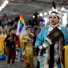 A group of people in native american costumes.|Two native americans standing next to each other.|A woman in colorful clothing and white arm bandages.|A woman with long black hair wearing a red shirt.