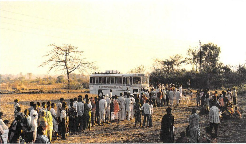 A group of people standing around a bus.|A group of people standing around in front of an ambulance.