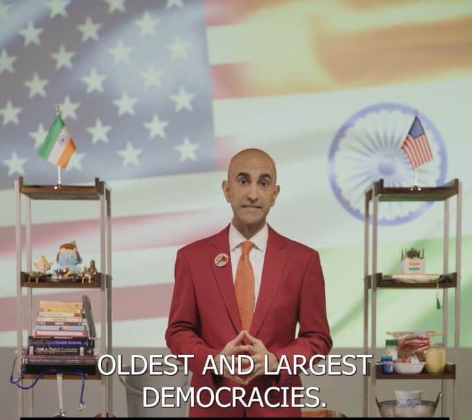 A man in red jacket standing next to two shelves.|A man in blue suit and tie holding a microphone.|A man in blue suit and tie holding a microphone.