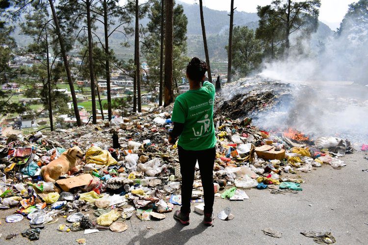 A person standing in front of garbage on the side of a road.|A truck is driving down the side of a river.|Two people picking up trash on a hill|A man and woman holding an award in front of some buildings.
