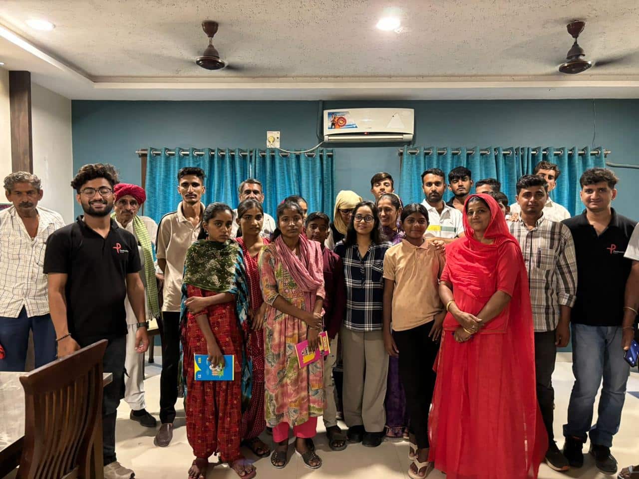 Community group posing in indoor meeting room|Woman with glasses speaking into microphone at conference|Group photo of smiling people in red room|Women with AIDS awareness banner and red ribbons
