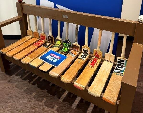 Bench made from cricket bats, colorful labels.|Benches made from cricket bats.|Person standing behind handcrafted wooden bench.