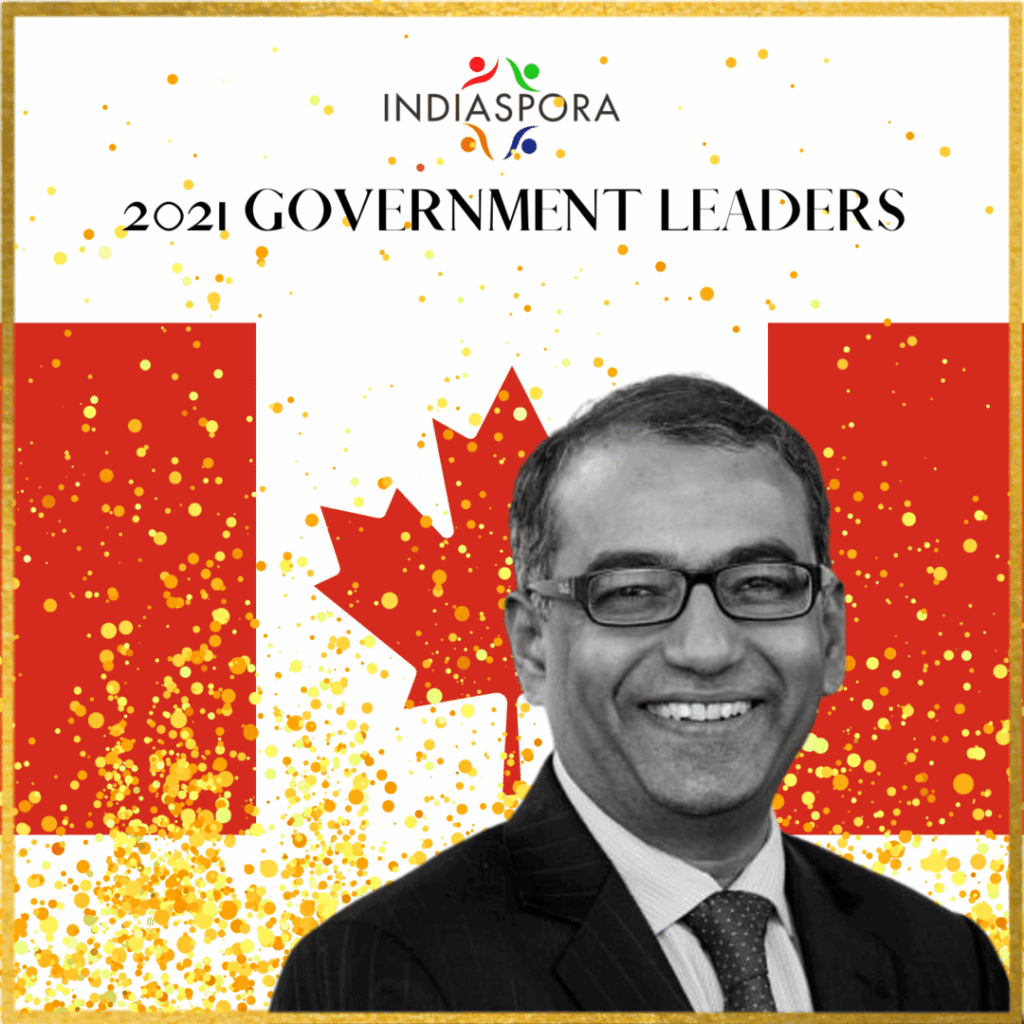 A man in glasses and suit standing next to canadian flag.|A man in glasses and suit standing next to canadian flag.