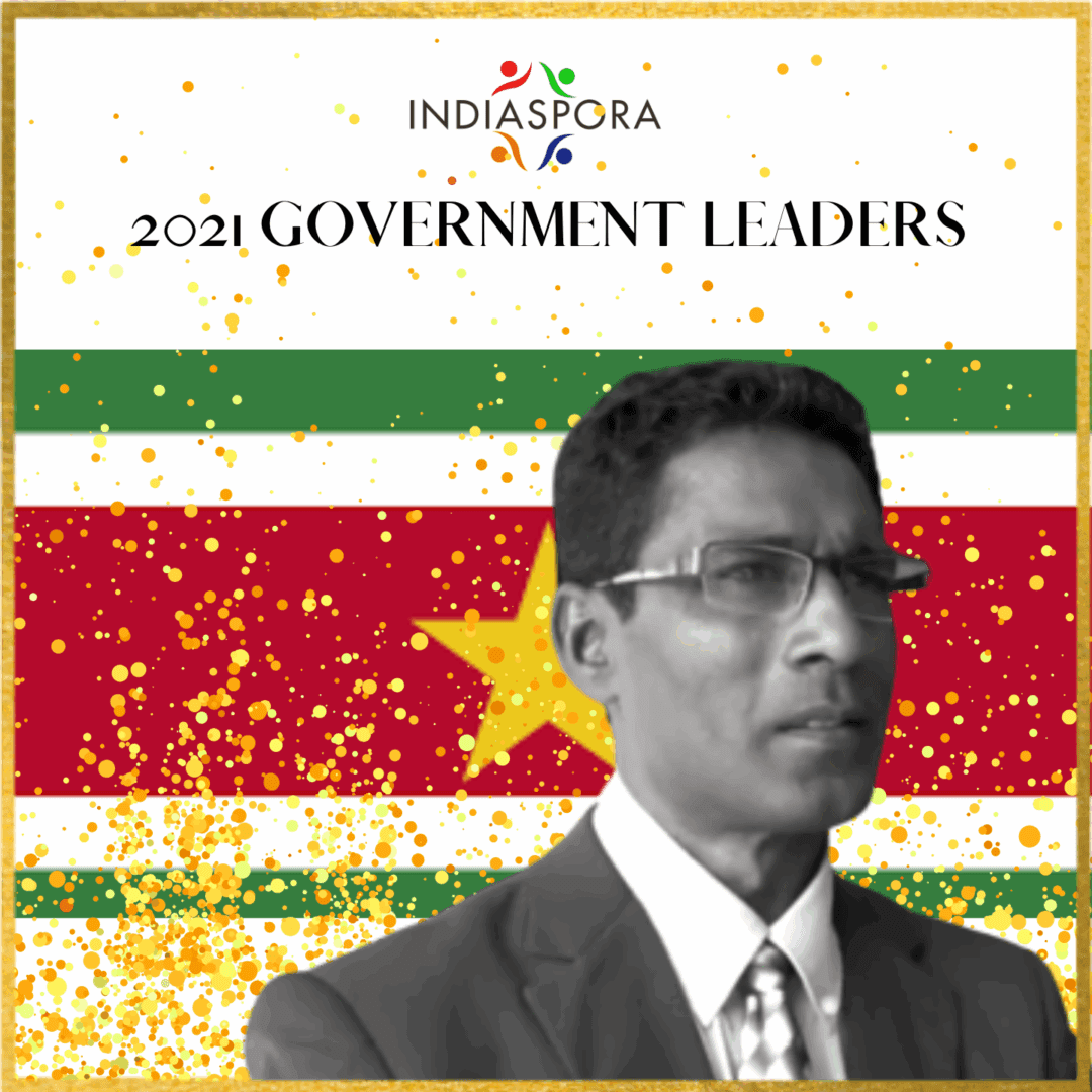 A man in glasses and suit standing next to the flag of suriname.