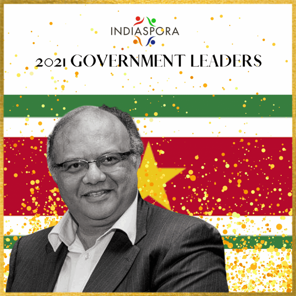 A man in glasses and suit standing next to the flag of suriname.