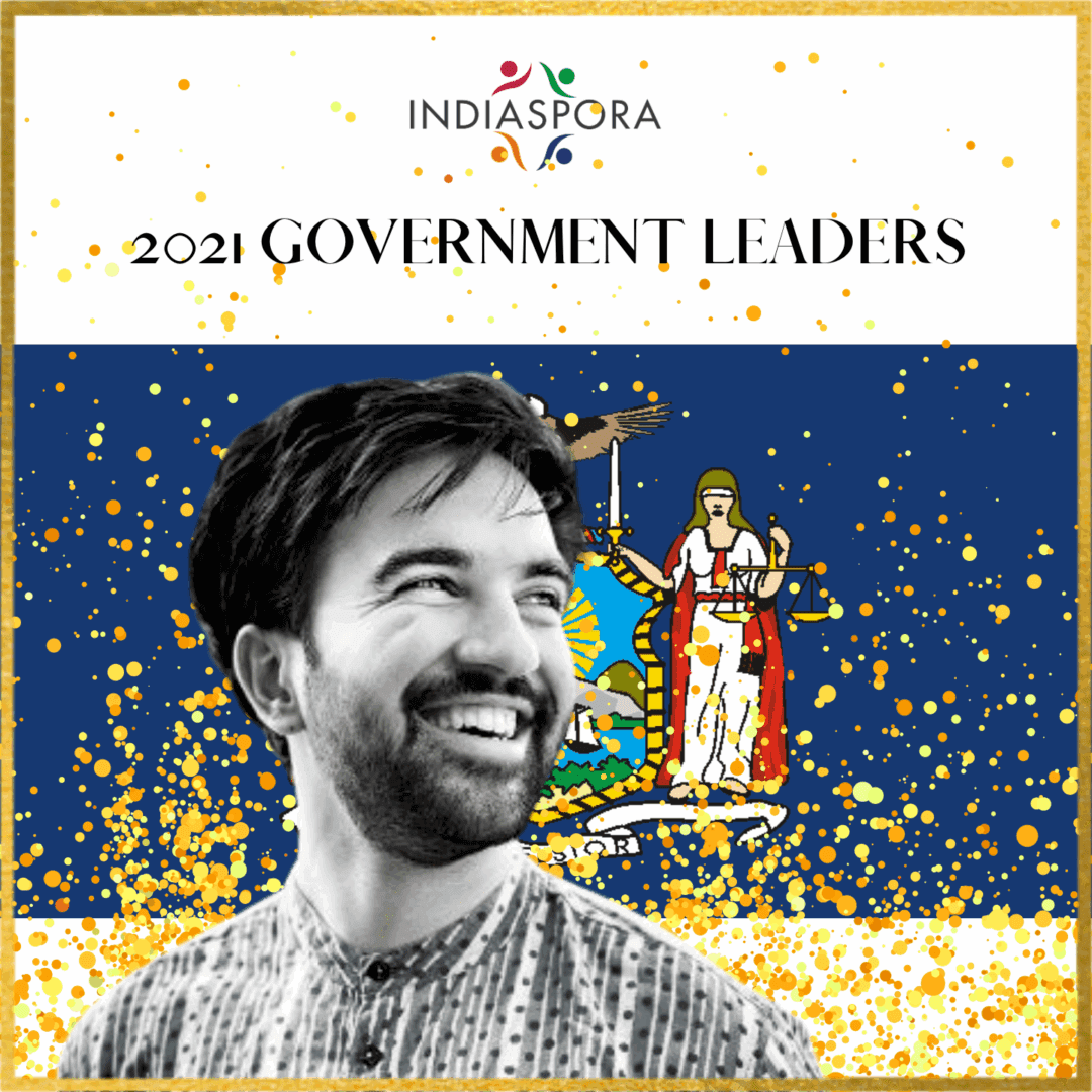 A man smiling in front of the state flag.